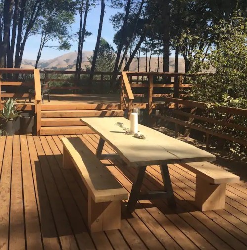 A wooden deck with a picnic table and benches, surrounded by trees and a scenic view in the background.