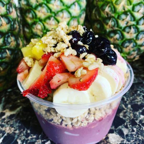 A colorful acai bowl topped with granola, strawberries, bananas, and blueberries, with pineapples in the background.