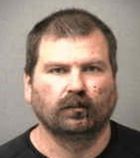 Mugshot of a man with short hair and a beard, wearing a black shirt, with visible facial marks.