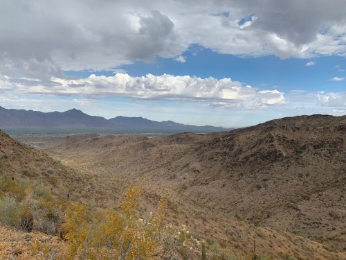 A panoramic view of a desert landscape with rolling hills, sparse vegetation, and a cloudy sky in the background.
