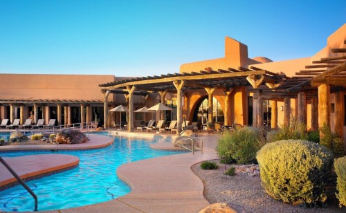 A serene pool area with lounge chairs, surrounded by desert landscaping and a modern adobe-style building under a clear blue sky.