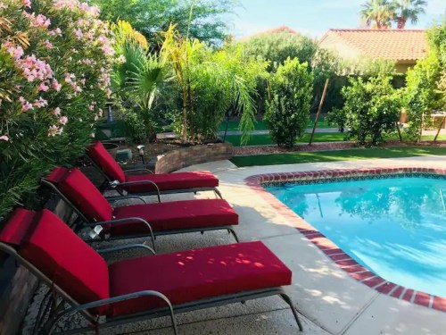 A serene backyard with a pool, surrounded by lush greenery and pink flowers, featuring red lounge chairs by the water.