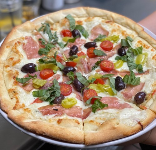 A freshly baked pizza topped with ham, cherry tomatoes, olives, peppers, and fresh basil on a thin crust.