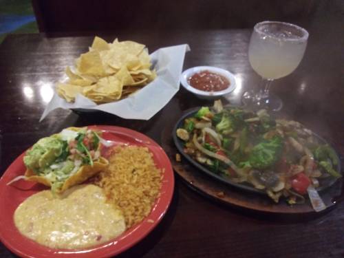 A plate with tacos, rice, and sizzling vegetables, accompanied by chips, salsa, and a margarita.