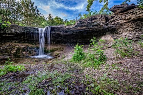 A serene waterfall cascading over rocky terrain, surrounded by lush greenery and a clear blue sky.
