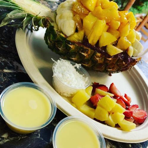 A pineapple filled with diced mango and banana, served with coconut, strawberries, and two cups of sauce.