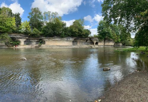 A serene river scene with rocky banks, lush greenery, and a partly cloudy sky reflecting on the water's surface.
