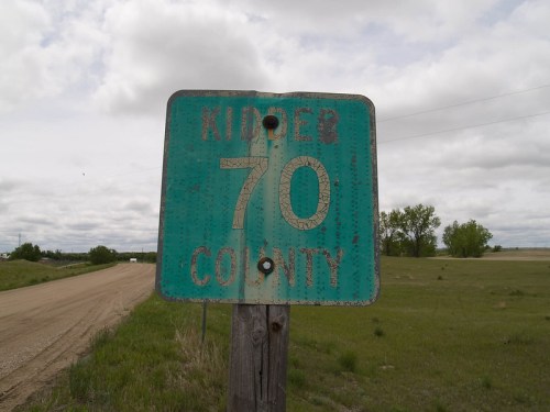 Weathered road sign for Kidder County, indicating the number 70, with a gravel road and cloudy sky in the background.