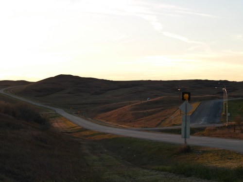 A rural road intersection at sunset, with rolling hills and a traffic signal in the foreground.