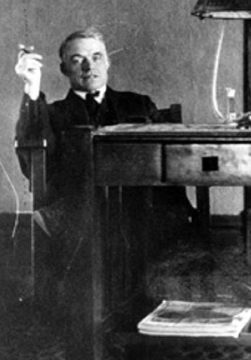 A man in formal attire sits at a wooden desk, holding a cigarette, with papers scattered nearby.