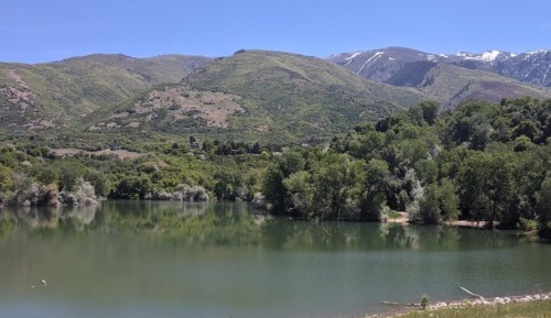 A serene lake surrounded by lush greenery and mountains under a clear blue sky. Snow-capped peaks in the distance.