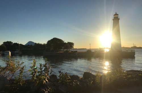 A lighthouse stands by the water at sunset, with trees and a mountain in the background, casting a warm glow.