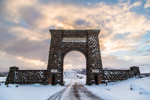 Stone archway with a sign, surrounded by snow and mountains under a cloudy sky.