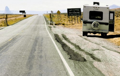 An RV parked beside a highway with a "Welcome to Arizona" sign in the background and a cracked road surface.