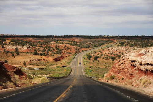 A winding road stretches through a desert landscape with red rock formations and green vegetation under a cloudy sky.