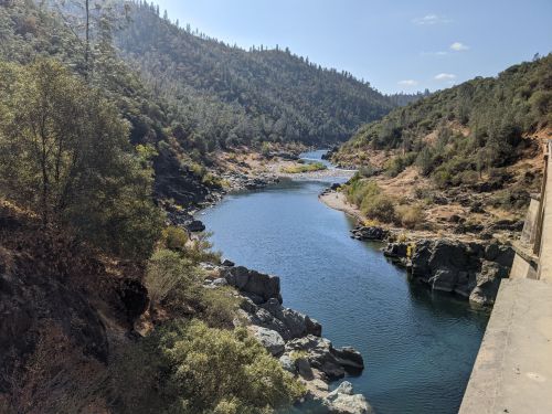 A serene river winding through a rocky landscape, surrounded by lush greenery and distant hills under a clear blue sky.