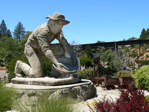 A large statue of a miner panning for gold, surrounded by greenery and historical mining equipment.