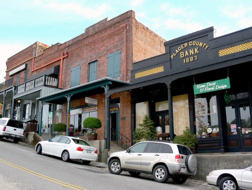 Historic buildings line a sloped street, featuring a bank from 1887 and various shops with greenery.