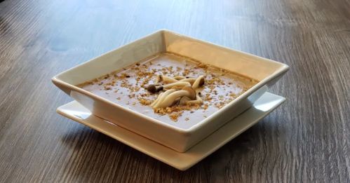 A square bowl of creamy soup topped with noodles and sprinkled with crushed nuts, placed on a wooden table.