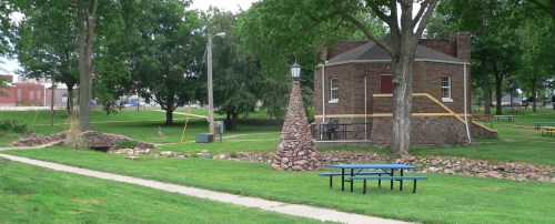 A park scene featuring a brick building, stone structures, and picnic tables surrounded by green grass and trees.