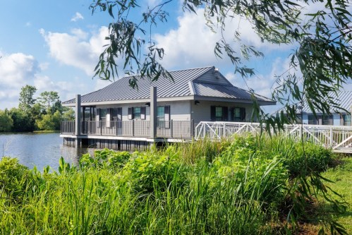 A modern building on stilts by a calm lake, surrounded by lush greenery and blue skies.