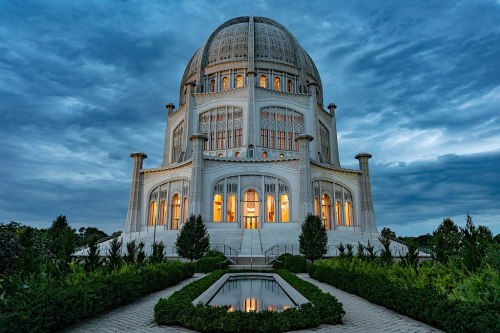 A grand white temple with a large dome, surrounded by greenery and reflecting water, under a dramatic blue sky.