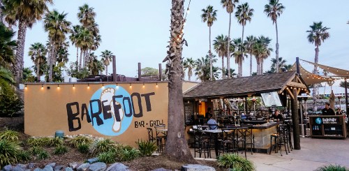 Exterior of Barefoot Bar & Grill, featuring palm trees, outdoor seating, and a vibrant mural on the wall.