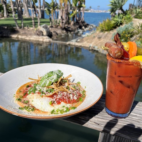 A plate of huevos rancheros with a colorful cocktail beside it, set against a scenic waterfront background.
