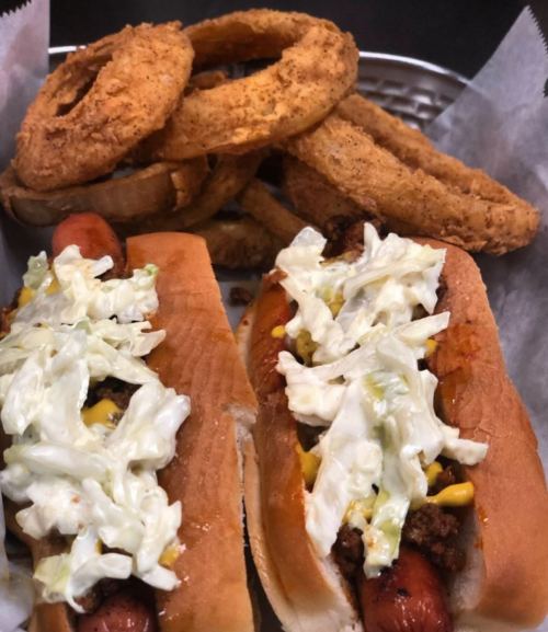 Two loaded hot dogs topped with coleslaw and mustard, served with a side of crispy onion rings.