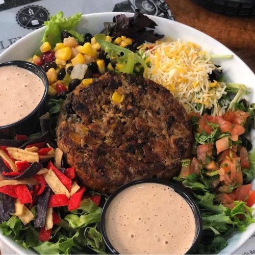 A colorful salad with a veggie patty, corn, black beans, cheese, and two cups of dressing on a bed of greens.