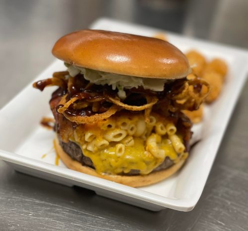 A close-up of a burger topped with macaroni and cheese, crispy onions, and barbecue sauce, served with side items.