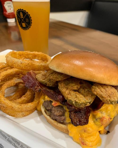 A hearty burger topped with bacon and fried pickles, served with onion rings and a glass of beer.