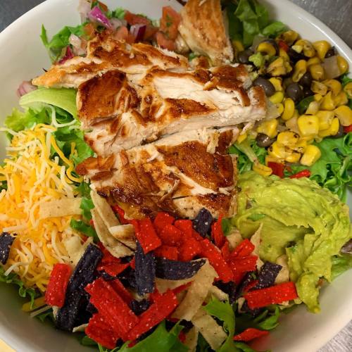 A colorful salad bowl with grilled chicken, lettuce, cheese, guacamole, corn, black beans, and tortilla strips.