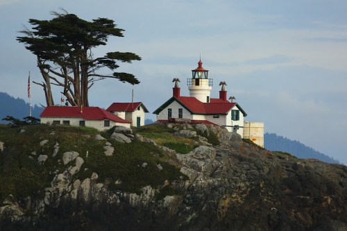 A scenic lighthouse on a rocky cliff, surrounded by trees and mountains in the background, with a cloudy sky.