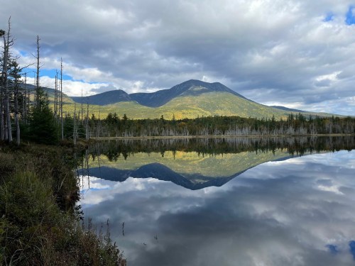 A serene lake reflects a mountain under a cloudy sky, surrounded by trees and autumn foliage.
