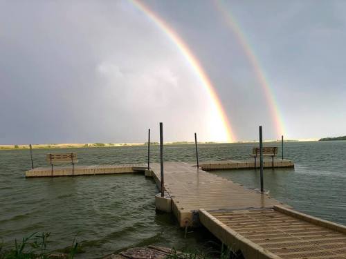 A wooden dock extends over a lake, with a double rainbow arching in the cloudy sky above.