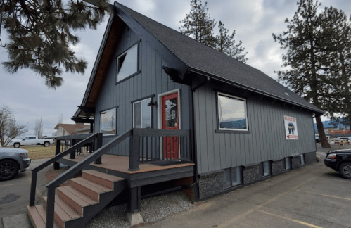 A gray, two-story building with a steep roof, wooden steps, and a red door, surrounded by trees and parked cars.