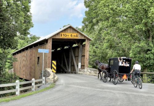 A horse-drawn carriage passes under a wooden covered bridge, with a cyclist nearby and trees in the background.