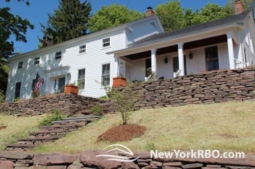 A white two-story house with a porch, surrounded by greenery and stone landscaping. An American flag is visible.