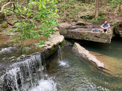 A person sits on a rock by a small waterfall in a lush, green forest. Water flows into a serene pool below.