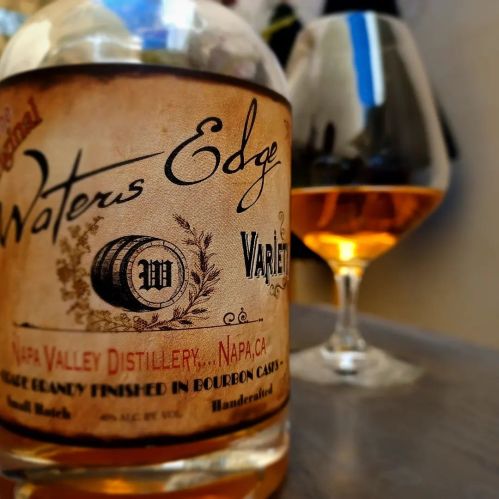 A close-up of a bottle labeled "Waters Edge" from Napa Valley Distillery, with a glass of amber liquid in the background.