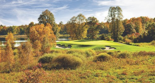 A scenic golf course surrounded by autumn foliage and a calm lake under a clear blue sky.