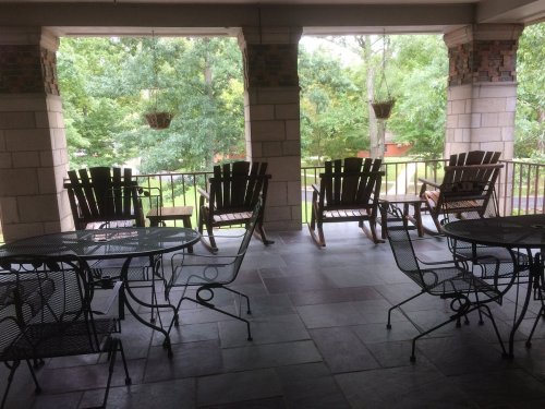 A covered porch with wooden rocking chairs and metal tables, surrounded by greenery and trees.