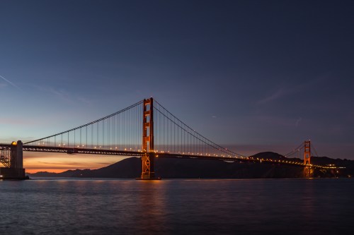 Golden Gate Bridge illuminated at dusk, spanning a calm bay with mountains in the background.