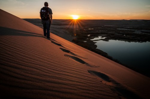 A person walks along a sandy dune at sunset, with footprints leading to a calm body of water in the background.