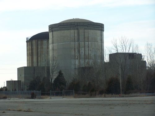 Two large, cylindrical concrete structures of a nuclear power plant, surrounded by barren land and sparse trees.