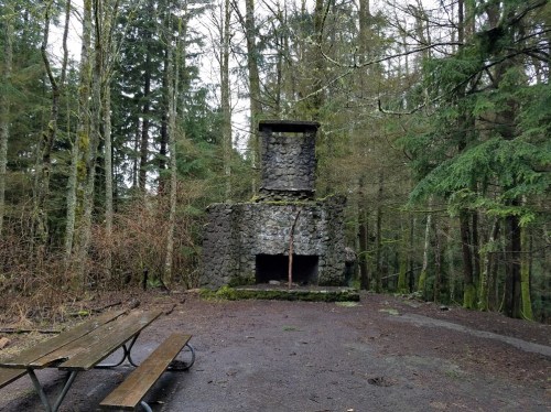 A stone fireplace stands in a forest clearing, surrounded by trees and a picnic table nearby.
