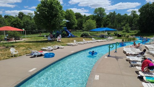 A sunny pool area with lounge chairs, a lazy river, and water slides surrounded by green trees and blue skies.