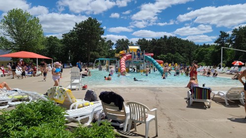 A busy outdoor pool area with children playing on slides, adults relaxing on lounge chairs, and a clear blue sky.