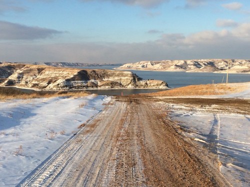 A dirt road leads down to a snowy landscape with hills and a lake under a partly cloudy sky.